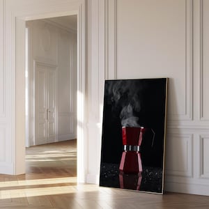 Puede incluir: Impresión enmarcada de una cafetera italiana roja con vapor saliendo de la boquilla. La cafetera está sobre un fondo negro, con un marco dorado. La imagen está apoyada contra una pared blanca.