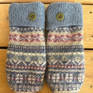 May include: A pair of knit mittens featuring a multi-coloured Fair Isle pattern in shades of blue, cream, and burgundy. Each mitten has a solid light blue ribbed cuff accented with a single round olive green button with a small pink centre.