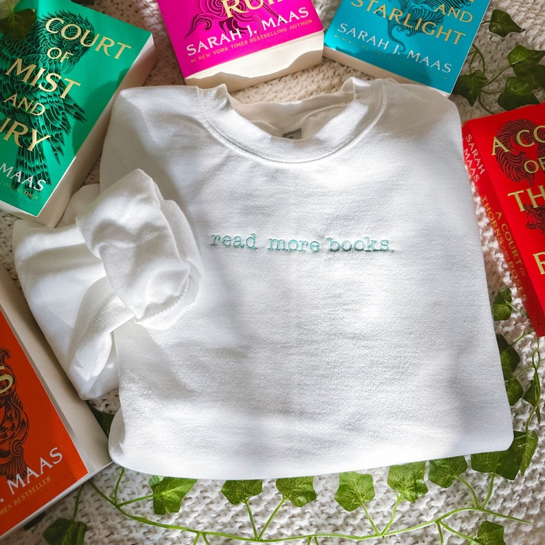 May include: White sweatshirt with the text "read more books." embroidered in light blue. The sweatshirt is surrounded by several books with colorful covers.