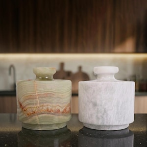 May include: Two decorative stone jars. One is green and brown, the other is white. Both have a cylindrical body and a small, round top. The jars are sitting on a dark surface.