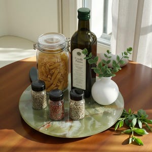 May include: A still-life arrangement featuring a green marble tray holding various kitchen items. Includes small spice jars, a glass jar of pasta, an olive oil bottle, and a small white vase with greenery. The items are arranged on a wooden table.