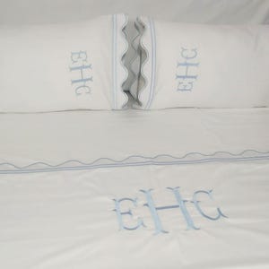May include: White bed linen set with light blue embroidered initials "EHC" on the pillowcases and flat sheet. The pillowcases have a scalloped edge with light blue trim. The flat sheet also features a light blue embroidered border.