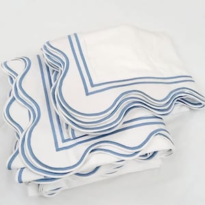 May include: White bedding set with blue scalloped edges and blue trim. The set includes a folded flat sheet and a folded fitted sheet. The sheets are made of a soft, smooth fabric.