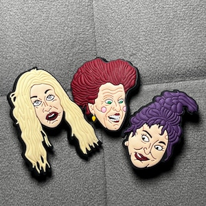 May include: Three colorful shoe charms featuring the Sanderson sisters from the movie Hocus Pocus. Each charm depicts a different sister's face with distinct hair colors: blonde, red, and purple. The charms have black borders.