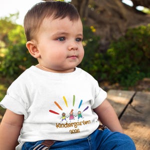 Könnte beinhalten: Ein weißes Kurzarm-T-Shirt mit einer bunten Grafik von drei Strichmännchen, die sich unter einem regenbogenfarbenen Sonnenstrahlen-Design an den Händen halten. Darunter steht der Text Kindergarten 2026 in brauner Schrift.