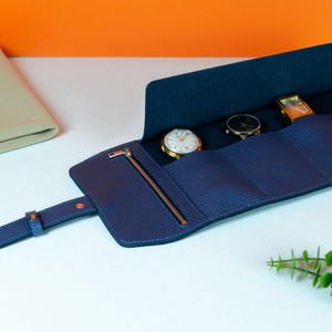 May include: A navy blue leather watch roll with a zipper closure and a strap. The roll is open, revealing three watches with gold and black faces. The background is white and orange.