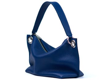 May include: A deep blue handbag with a single shoulder strap. The bag features a textured surface, a silver zipper, and silver metal rings attaching the strap to the bag. The bag is set against a white background.