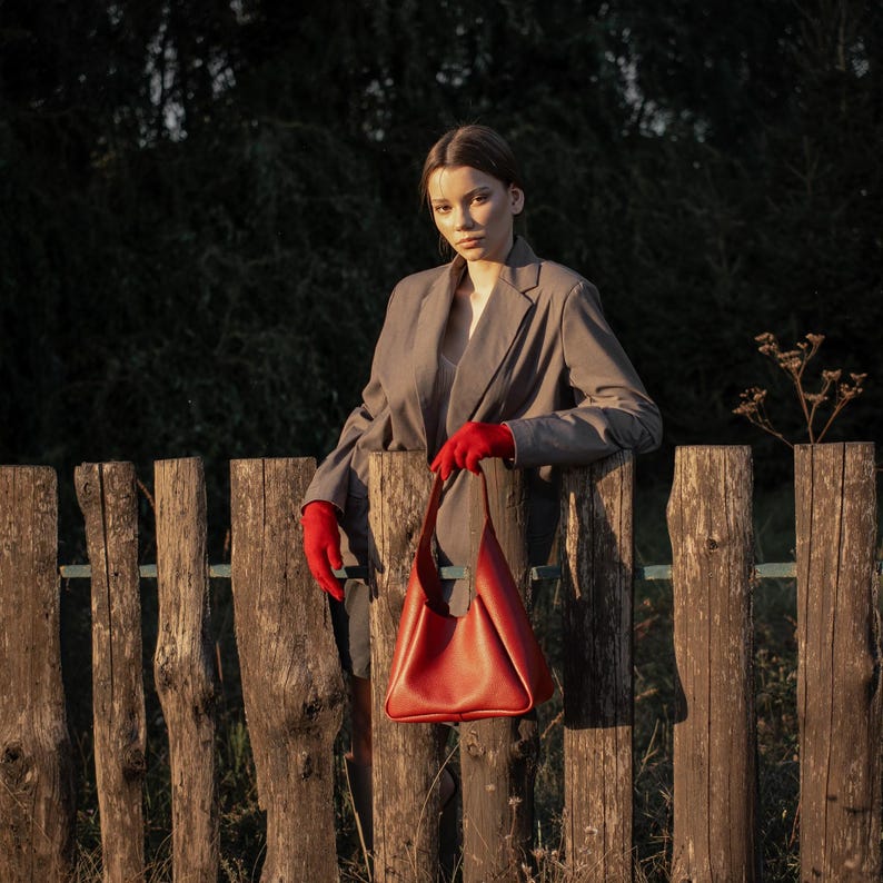 Woman standing by wooden fence holding red leather hobo bag
