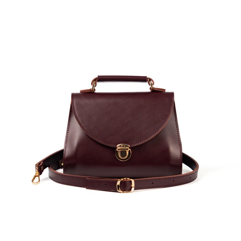 Burgundy leather top handle bag with gold clasp and detachable crossbody strap on white background.