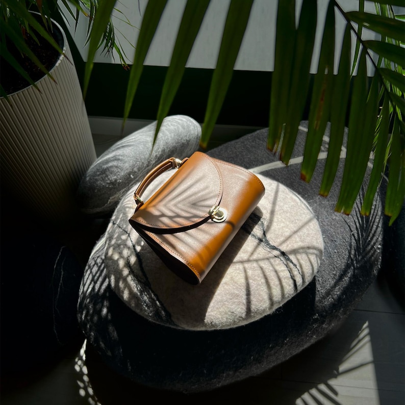 Brown leather handbag with handle and clasp placed on stone decor with sunlight shadows.
