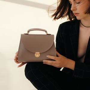 Woman in dark suit holding gray leather crossbody bag in soft light.