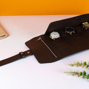 May include: A brown leather watch roll with three watches inside. The watches have gold and black watch faces. The watch roll has a zipper pocket and a strap with a gold buckle. The background is white and orange.