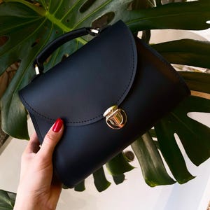 Black leather handbag with gold clasp held in hand over green monstera leaves.
