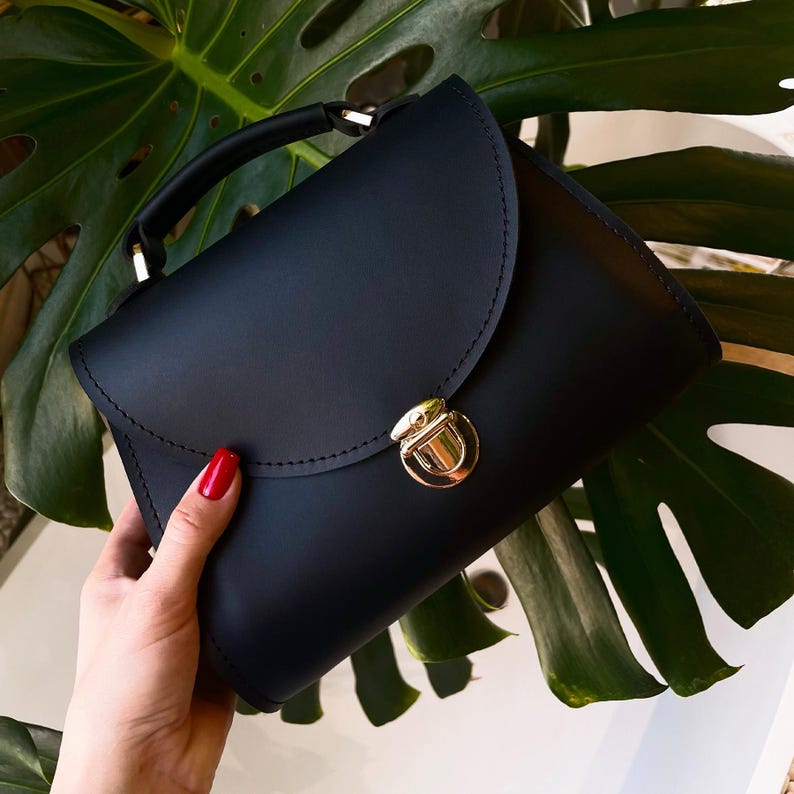 Black leather handbag with gold clasp held in hand over green monstera leaves.