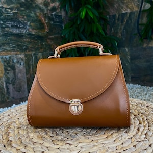 Brown structured leather handbag on woven surface with green plants background.