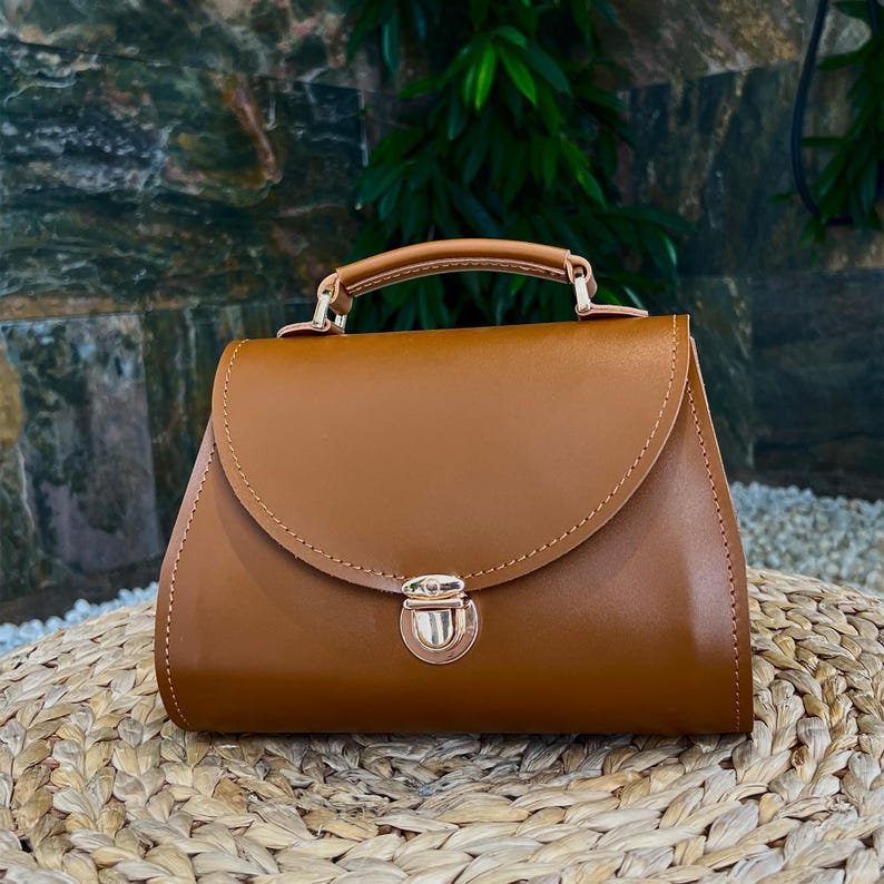 Brown structured leather handbag on woven surface with green plants background.