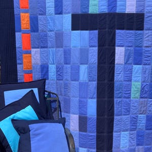 Peut inclure: Une courtepointe en patchwork bleu et bleu marine avec un grand 'T' bleu marine au centre. La courtepointe est entourée de quatre coussins bleus et bleu marine.