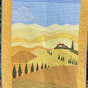 Peut inclure: Une tenture murale matelassée avec un motif de paysage. Le fond est un ciel bleu avec un nuage blanc. Au premier plan, une série de collines jaunes, brunes et vertes avec une petite maison rouge sur une colline. Il y a plusieurs arbres vert foncé sur les collines.