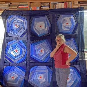 Op de afbeelding: Een blauw en wit quilt met een geometrisch patroon van concentrische cirkels. De quilt hangt aan een muur en een persoon staat er voor.