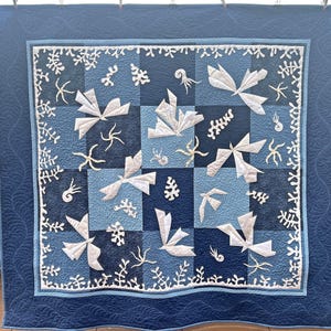 May include: A square quilt featuring a checkerboard pattern of dark and light blue fabric squares. The quilt is decorated with white fabric appliques of stylized birds, coral, and spiral shells, finished with a dark blue border with stitching.