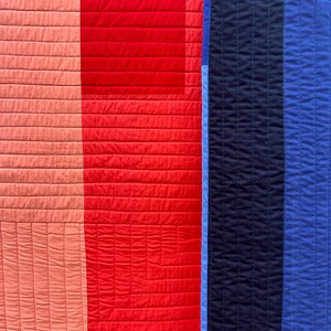 Op de afbeelding: Een textielquilt met verticale kleurvlakken in tinten koraal, felrood, lichtblauw, marineblauw en koningsblauw. De stof is over het gehele oppervlak doorgestikt met een consistent horizontaal en verticaal rasterpatroon.