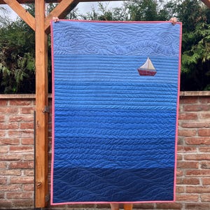 Peut inclure: Une courtepointe rectangulaire avec un motif bleu dégradé passant de teintes claires à foncées. Elle présente une applique de voilier et le texte brodé, May the wind be at your back. L'ensemble est bordé d'une bordure rose corail.
