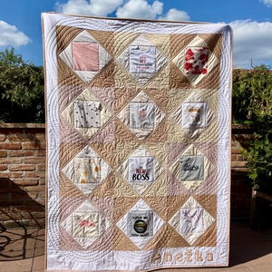 Peut inclure: Une courtepointe souvenir rectangulaire composée d'une grille de douze panneaux carrés en tissu provenant de divers vêtements de bébé, avec des motifs et du texte comme "THE NEW BOSS" et "LOVE ME YOU MUST". Bordures matelassées beige et blanc.