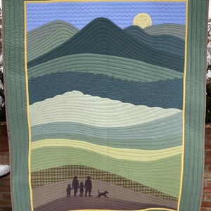 May include: A green quilted wall hanging with a landscape design. The design features a family of three and a dog walking in front of a mountain range with a yellow sun in the sky.