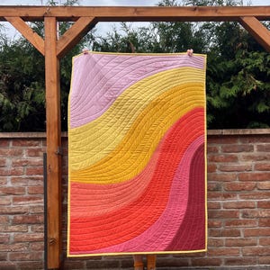 Op de afbeelding: Een rechthoekige quilt met golvende, verticale kleurstroken in lichtroze, geel, oranje, rood en donkerroze. De stof is doorgestikt met gebogen, parallelle lijnen en heeft een gele rand. De tekst "have a good day" is erin gestikt.