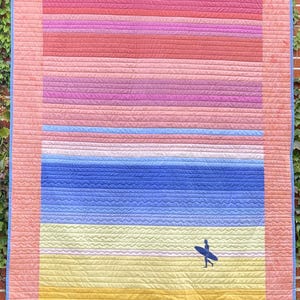 Peut inclure: Une tenture murale matelassée avec une scène de plage. La courtepointe présente des bandes horizontales de couleurs, notamment rose, rouge, violet, bleu et jaune. Une silhouette d'une personne avec une planche de surf est sur la plage. La courtepointe a une bordure rose.