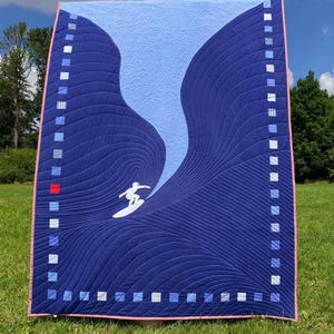 Peut inclure: Une courtepointe bleue et bleu clair avec une silhouette blanche d'un surfeur chevauchant une vague. La courtepointe est matelassée avec un motif tourbillonnant et a une bordure rose.