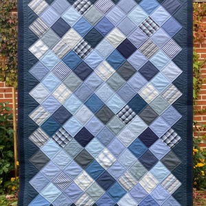 Könnte beinhalten: Eine blaue und weiße Quiltdecke aus wiederverwendetem Stoff. Die Decke besteht aus Quadraten aus verschiedenen Blautönen und weißem Stoff, die in einem Diamantenmuster angeordnet sind. Die Decke ist mit einem dunkelblauen Stoff eingefasst.