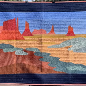 Peut inclure: Une courtepointe rectangulaire représentant un paysage désertique avec des formations rocheuses rouges sur un ciel bleu clair. Le premier plan présente des pièces de tissu vert sauge, beige et terre cuite, bordées d'une large bordure bleu marine.