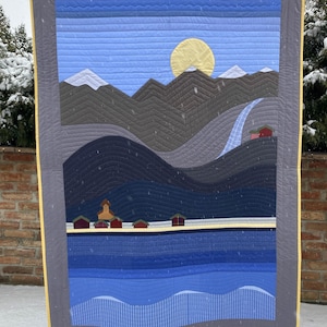 Op de afbeelding: Een landschapskwilt met een berglandschap met een gele zon, blauwe lucht en water. De quilt gebruikt tinten blauw, grijs en bruin, met kleine rode gebouwen langs de waterkant. De quilt is omlijst met een grijze rand.