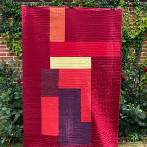 May include: A red quilt with geometric shapes in shades of red, orange, purple, and yellow. The quilt is hanging on a clothesline in front of a brick wall covered in ivy.