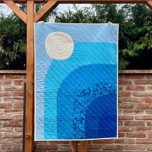 Peut inclure: Une courtepointe rectangulaire présentant un dégradé de nuances bleues disposées en motifs d'arcs concentriques. Un grand motif de soleil circulaire de couleur crème avec un matelassage en spirale est situé dans le coin supérieur gauche.
