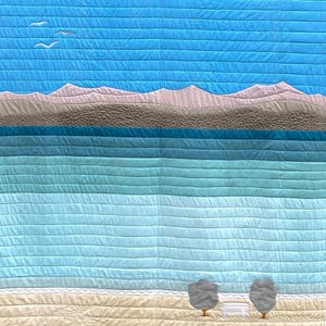 Op de afbeelding: Een gestikte wandhanger met een strandscène. De scène toont een blauwe lucht met twee witte vogels die vliegen, een rij lichtroze bergen, een blauwe zee met lichtblauwe golven en een beige strand met twee grijze bomen en een witte bank.