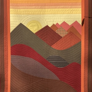 May include: A quilted wall hanging with a geometric design of mountains and a sunset. The mountains are in shades of brown, green, and red, and the sunset is in shades of pink, orange, and yellow.