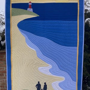 Op de afbeelding: Een gestikte wandhanger met een strandscene. De scène toont een vuurtoren op een klein eiland, een zandstrand en een blauwe zee met golven. Een gezin van drie loopt in silhouet over het strand.