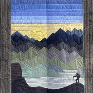 Könnte beinhalten: Ein gesteppter Wandbehang mit einer Bergszene. Die Berge bestehen aus verschiedenen Blautönen und Grüntönen. Eine gelbe Sonne geht hinter den Bergen unter. Eine Silhouette eines Wanderers steht auf einem Berggipfel.