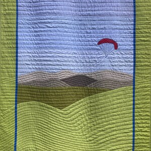 Op de afbeelding: Een groene en blauwe gequilte wandhanger met een rode en witte parachute in de lucht boven een landschap met bruine heuvels.