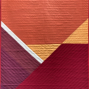 Op de afbeelding: Een quilt met een geometrisch ontwerp met een groot rood gedeelte, een kleiner paars gedeelte, een wit en geel gedeelte en een klein oranje gedeelte. De quilt is gestikt met diagonale lijnen.