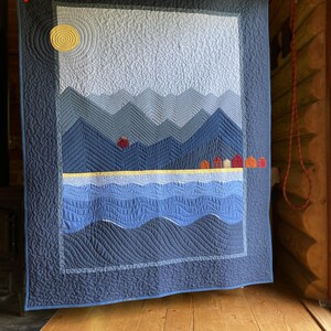 May include: A handmade quilt showing a landscape scene with mountains, water, and a sun. The quilt uses shades of blue, grey, yellow, and red. The design includes a sun in the upper left corner and a row of small houses along the horizon.