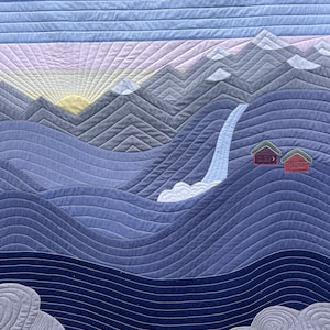 以下が含まれることがあります： 滝、小さな家2軒、手前に赤いカヌーのある山岳風景を描いたキルト。空は青、ピンク、黄色のグラデーションで、山々の上に昇る太陽が描かれている。キルトは様々な青色の生地で作られている。