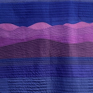 Peut inclure: Une courtepointe de couleurs bleu et violet. La courtepointe est faite de différents tissus et présente un motif matelassé. La courtepointe est composée de bandes horizontales de différentes nuances de bleu et de violet. La bande supérieure est bleu clair avec des pois blancs. La bande suivante est bleu foncé. La bande suivante est violet clair avec un motif ondulé. La bande suivante est violet foncé. La bande suivante est bleu clair. La bande inférieure est bleu foncé avec des pois blancs.