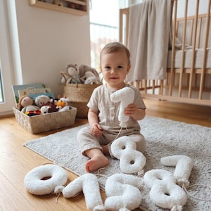 May include: A set of white, plush, textured fabric numbers shaped like balloons, each featuring a small tied knot at the base with a thin string attached. The numbers are scattered on a gray rug, with one number held up by a child in a beige outfit.