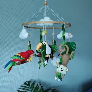 May include: A baby mobile featuring a wooden ring with felt clouds, a red parrot, a toucan, and a monkey. The mobile is suspended by white strings and includes green leaves and white flowers, creating a jungle theme.