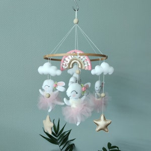 May include: A baby mobile with a pink and gold rainbow, white clouds, and three white bunny toys wearing pink tutus. The mobile also has two gold stars.