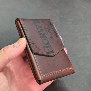 May include: A dark brown leather card holder with a flap closure and visible stitching. The front features a black stamped logo that reads HORWEEN FINEST QUALITY LEATHER SINCE 1905, with a central emblem depicting a horse and a rider.