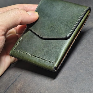 Op de afbeelding: Een bosgroene lederen kaarthouder met een puntige klepsluiting en zichtbare donkere stiksels aan de zijkant. De portemonnee heeft een gladde, matte textuur en een gestructureerd, compact ontwerp met strakke randen en een functionele uitstraling.